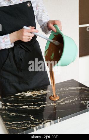Nahaufnahme - Frau rührt Schokolade und gießt auf einem Steinbrett für die Herstellung von hausgemachten Pralinen. Süßwarenherstellung, Konditorei, Dessertkonzept Stockfoto