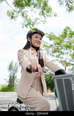 Lächelnde junge asiatische Geschäftsfrau in beigefarbenem Anzug, die mit dem Fahrrad ins Büro fährt Stockfoto