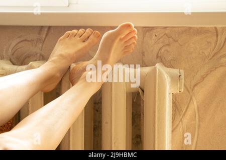 Die Füße der Frauen liegen auf einer alten weißen gusseisernen Batterie in einer Wohnung zu Hause, einem warmen Haus im Winter, einer Heizperiode Stockfoto