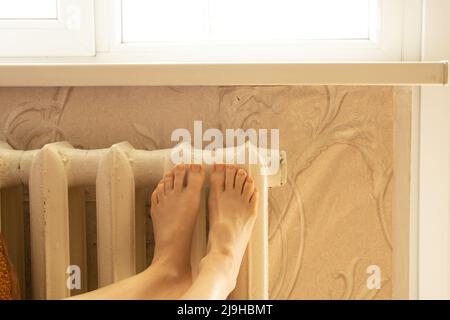 Die Füße der Frauen liegen auf einer alten weißen gusseisernen Batterie in einer Wohnung zu Hause, einem warmen Haus im Winter, einer Heizperiode Stockfoto