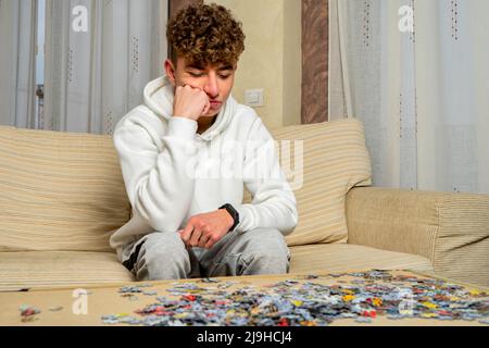 Ein junger Mann mit lockigen Haaren sitzt zu Hause auf dem Sofa und versucht, ein Puzzle aus vielen Teilen zusammenzubauen Stockfoto