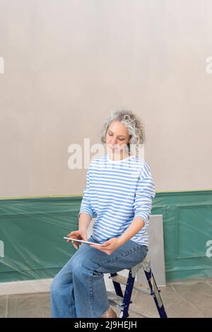 Frau, die zu Hause auf einer Leiter mit einem Tablet-PC vor der Wand sitzt Stockfoto
