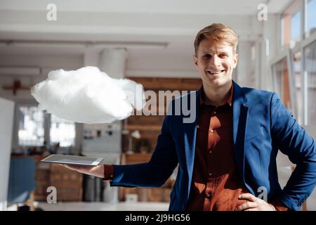 Glücklicher Geschäftsmann hält Tablet-PC unter schwebendem Cloud-Netzwerk im Büro Stockfoto