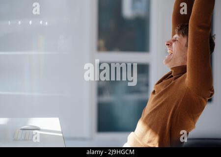 Ein glücklicher Geschäftsmann mit erhobenen Armen sitzt im Büro am Schreibtisch Stockfoto