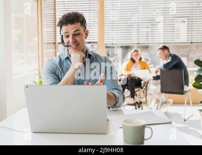 Junger Mann mit Headset und Laptop am Schreibtisch im Büro Stockfoto