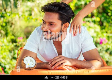 indischer Mann mit Öl Ayurveda Spa Treatment.Handsome bärtigen dunkelhaarigen männlichen Modell entspannen am Beach.Spa und Entspannung Konzept plumeria und Kokosnuss Stockfoto