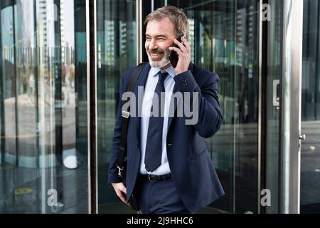 Lächelnder, reifer Geschäftsmann, der mit einem Smartphone spricht und aus der Drehtür herauskommt Stockfoto