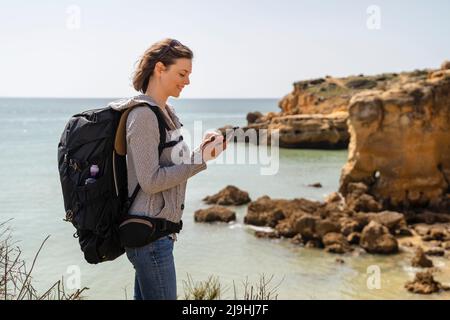 Lächelnde Frau mit Rucksack und Smartphone, die vor dem Meer steht Stockfoto