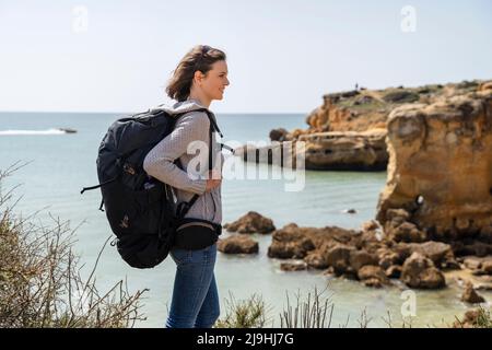 Lächelnde Frau mit Rucksack, die an sonnigen Tagen vor dem Meer steht Stockfoto