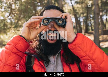 Fröhlicher bärtiger Mann mit offenem Mund und Blick durch ein Fernglas im Wald Stockfoto