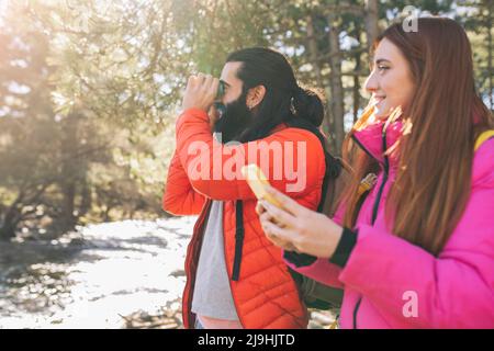 Lächelnder bärtiger Mann, der durch ein Fernglas schaut und bei einer Frau im Wald steht Stockfoto