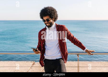 Glücklicher Mann, der an einem sonnigen Tag mit dem Mobiltelefon vor dem Geländer steht Stockfoto
