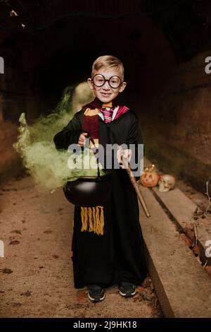 Lächelnder Junge, der im Tunnel einen Zauberkessel mit grünem Rauch hält und ein Kostüm trägt Stockfoto