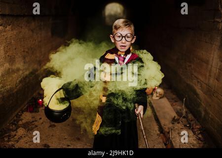 Netter Junge mit Kessel und Zauberstab, der inmitten des gelben Rauchs im Tunnel steht Stockfoto