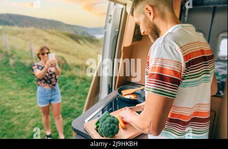 Junger Mann kocht Gemüse in einem Wohnmobil, während seine Frau mit ihrem Handy ein Foto von ihm macht Stockfoto