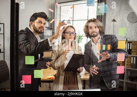 Team aus drei verschiedenen Mitarbeitern in formeller Kleidung mit Klemmbrett und Tablet, während sie vor einer Glaswand im Bürobereich stehen. Intelligente Kollegen, die mit neuen Ideen für ein gemeinsames Projekt austauschen. Stockfoto