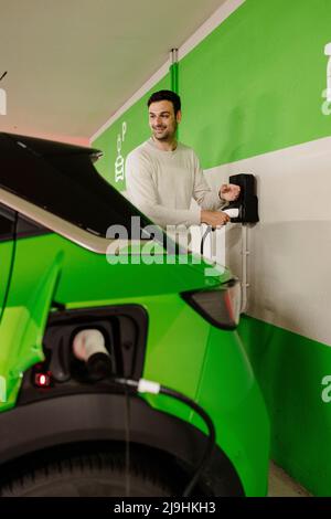 Glücklicher Mann lädt grünes Elektroauto an der Ladestation Stockfoto