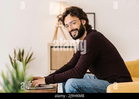 Lächelnder bärtiger Hipster mit Laptop, der zu Hause auf dem Sofa sitzt Stockfoto