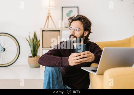 Nachdenklicher bärtiger Mann mit Mobiltelefon und Laptop, der zu Hause am Sofa sitzt Stockfoto
