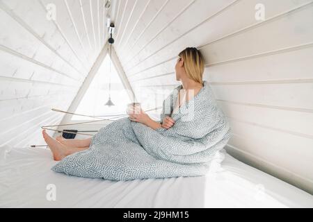 Frau mit Decke bedeckt, die zu Hause auf dem Dachboden auf dem Bett sitzt Stockfoto