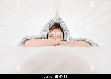 Verängstigte Frau, die unter der Decke auf dem Bett auf dem Dachboden liegt Stockfoto