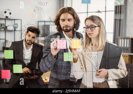Ein paar Arbeiter stehen vor einer Glaswand und schreiben Ideen auf Haftnotizen mit einem indischen männlichen Kollegen auf dem Hintergrund. Junge Mitarbeiter in formellen Wear Sharing Erfahrungen während des Bürotreffens. Stockfoto
