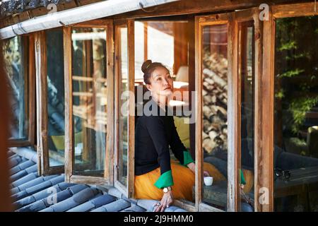 asiatische Frau, die auf der Fensterbank sitzt und eine Tasse Kaffee genießt Stockfoto