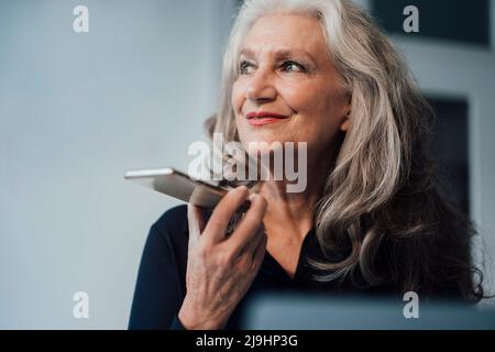 Lächelnde Geschäftsfrau, die über einen Sprecher im Büro auf dem Smartphone spricht Stockfoto