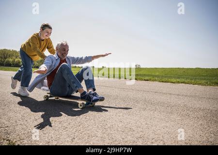 Verspielte Enkelin schiebt glücklichen Großvater auf Skateboard sitzen Stockfoto
