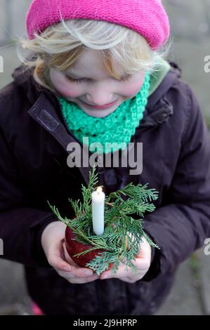 Lächelndes Mädchen, das mit Tannennadeln die brennende Gedächtniskerze auf dem Apfel anschaut Stockfoto