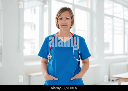 Lächelnder Arzt, der mit Händen in blauen Peelings-Taschen im Krankenhaus steht Stockfoto
