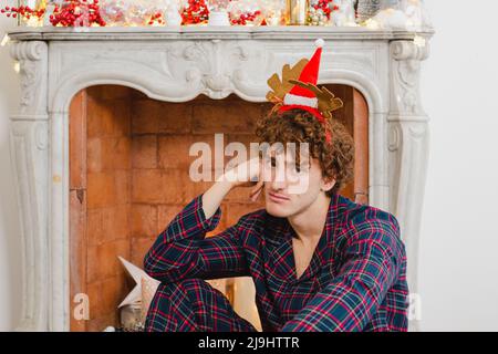 Gelangweilter junger Mann mit weihnachtlicher Kopfbedeckung, der vor einem Marmorkamin sitzt Stockfoto