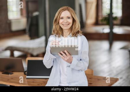 Lächelnde Geschäftsfrau mit Tablet-PC, der sich zu Hause auf den Tisch lehnt Stockfoto