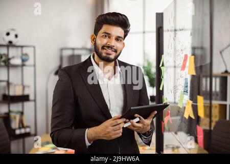 Porträt eines indischen Geschäftsführers, der formelle Kleidung trägt, ein digitales Tablet hält und neben einer Glaswand mit klebrigen Papieren steht. Konzept von Unternehmen, Menschen und Technologie. Stockfoto