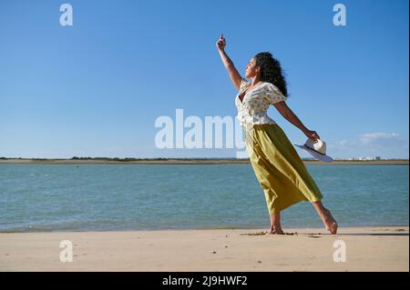Junge Frau mit erhobener Hand tanzt am Strand an der Küste Stockfoto