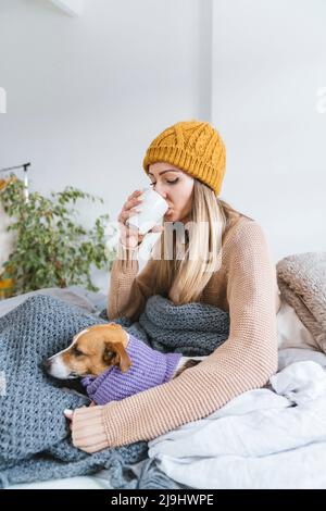 Junge Frau trägt wolligen Hut trinkenden heißen Tee im Bett mit Hund sitzen Stockfoto