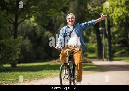 Ein glücklicher älterer Mann mit erhobenen Armen genießt eine Fahrradtour im Park Stockfoto