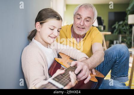 Lächelnder älterer Mann, der zu Hause der Enkelin Gitarre beibringt Stockfoto