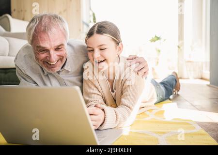 Glücklicher älterer Mann mit Enkelin, der einen Laptop benutzt und im Wohnzimmer zu Hause auf dem Teppich liegt Stockfoto