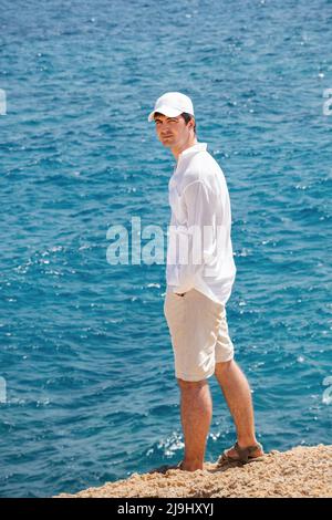 Rückansicht eines Mannes auf einem Hintergrund von gelbem felsigen Strand und blauem Meer. Ein Mann in einem weißen Leinenhemd, Shorts, Sandalen und einer Mütze. Reiseurlaub. Hände Stockfoto