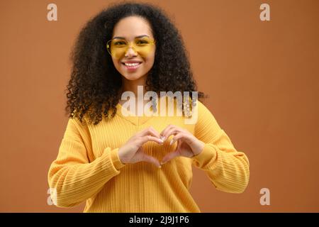 Lächelnd attraktive Brünette Frau schaut auf die Kamera und zeigt Herzform mit ihren Handflächen, gestikulende Liebe, romantisches Stimmungskonzept, isoliert auf Braun Stockfoto