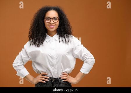 Waist up Porträt der fröhlichen weiblichen Youngster isoliert braunen Hintergrund. Junge multirassische Frau, die lächelnd lächelt, steht in der Nähe von leerem Raum Stockfoto