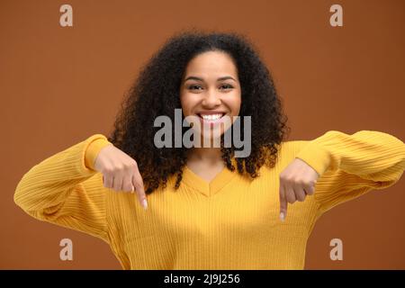 Charmante junge afroamerikanische Frau zeigt die Zeigefinger nach unten und fördert Neuheit isoliert auf braunem Hintergrund, Werbekonzept Stockfoto