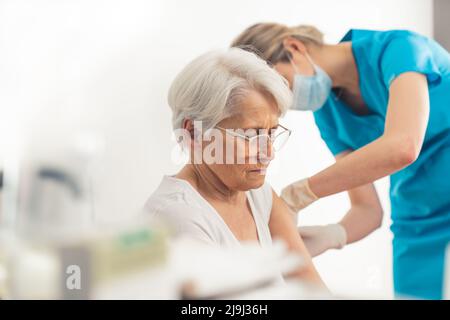 Blonde weibliche MD, die einer einschüchternden älteren Patientin einen Coronavirus-Impfstoff verabreicht. Innenaufnahme. Hochwertige Fotos Stockfoto