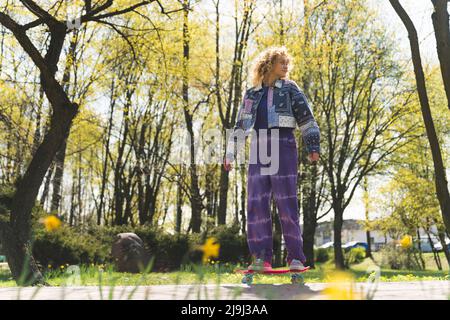 Blonde junge, große Mädchen mit einer Jeansjacke und einer lockeren Hose, die auf einem Skateboard steht und Spaß in einem Park hat. Hochwertige Fotos Stockfoto