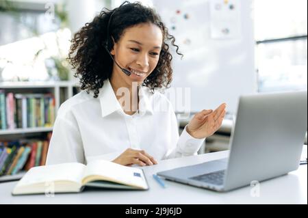 Erfolgreiche, smarte afroamerikanische Frau, Supportarbeiterin, Online-Consultant, Callcenter-Mitarbeiterin, sitzt an ihrem Schreibtisch vor einem Laptop, hat ein Video-Meeting mit einem Kunden, gestikuliert, lächelnd Stockfoto