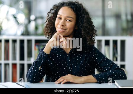 Porträt eines erfolgreichen jungen erwachsenen afroamerikanischen Mädchens, mit lockigen Haaren, Schüler, Lehrer, Mentor, In stilvoller Business-Kleidung sitzt man an einem Schreibtisch, blickt auf die Kamera, ruht sich von der Arbeit aus und lächelt freundlich Stockfoto