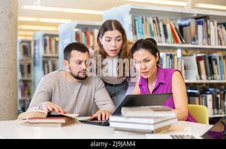 Drei Erwachsene Studenten arbeiten an einem Laptop und lesen Bücher in einer öffentlichen Bibliothek Stockfoto