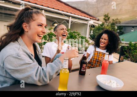 Lachen über kalte Getränke. Drei glückliche Freundinnen lachen fröhlich, während sie auf dem Dach zusammen hängen. Gruppe von jungen Freundinnen mit einem Stockfoto