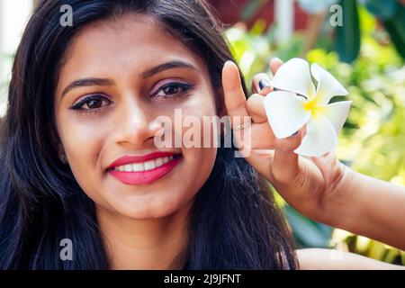 Nahaufnahme Kopfschuss Porträt der Haltung zuversichtlich lächelnd glücklich hübsche junge Frau Hintergrund von verschwommenen Bäumen, Plumeria Blumen. Positive menschliche Emotionen Stockfoto